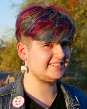 an individual with dyed hair in a denim jacket with a prominent pronoun pin displaying they, them, theirs